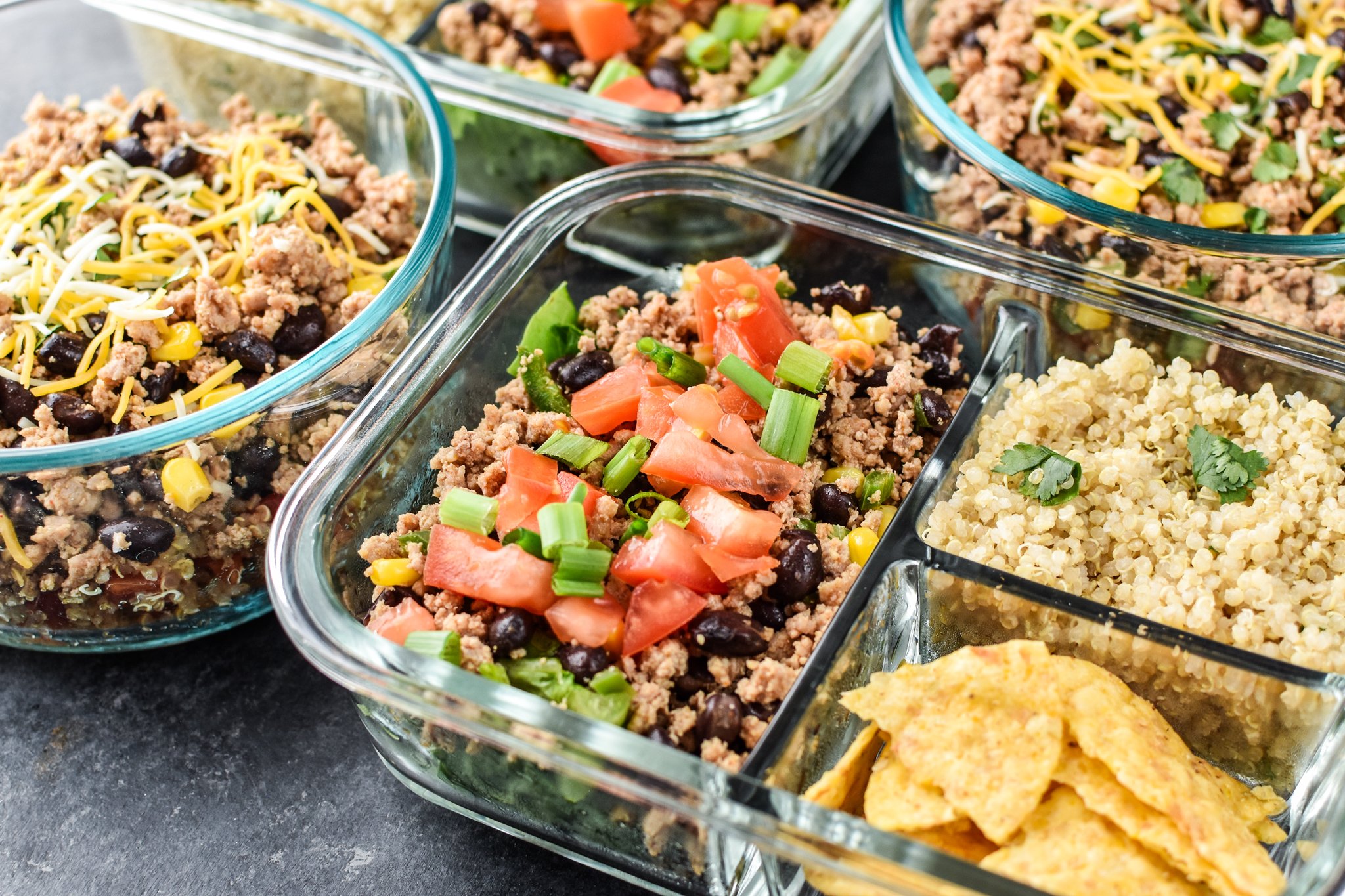 Four meal prepped lunches viewed from above for Hot & Cold Turkey Taco Meal Prep; two taco salads with quinoa and chips on the side and two taco bowls with tomato and green chili quinoa.