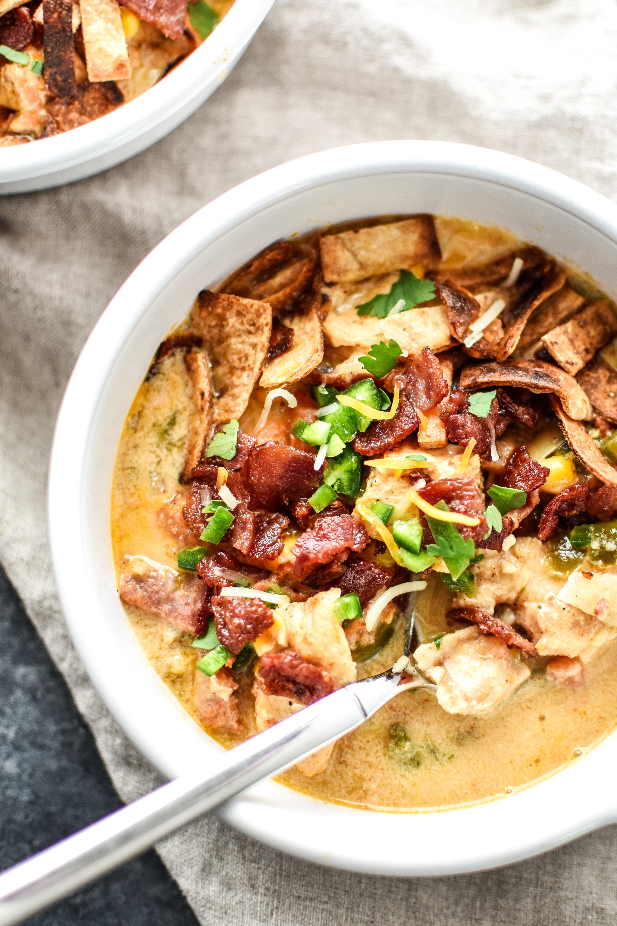 A bowl of jalapeno popper chicken soup with bacon bits, tortilla strips and shredded cheese on top.