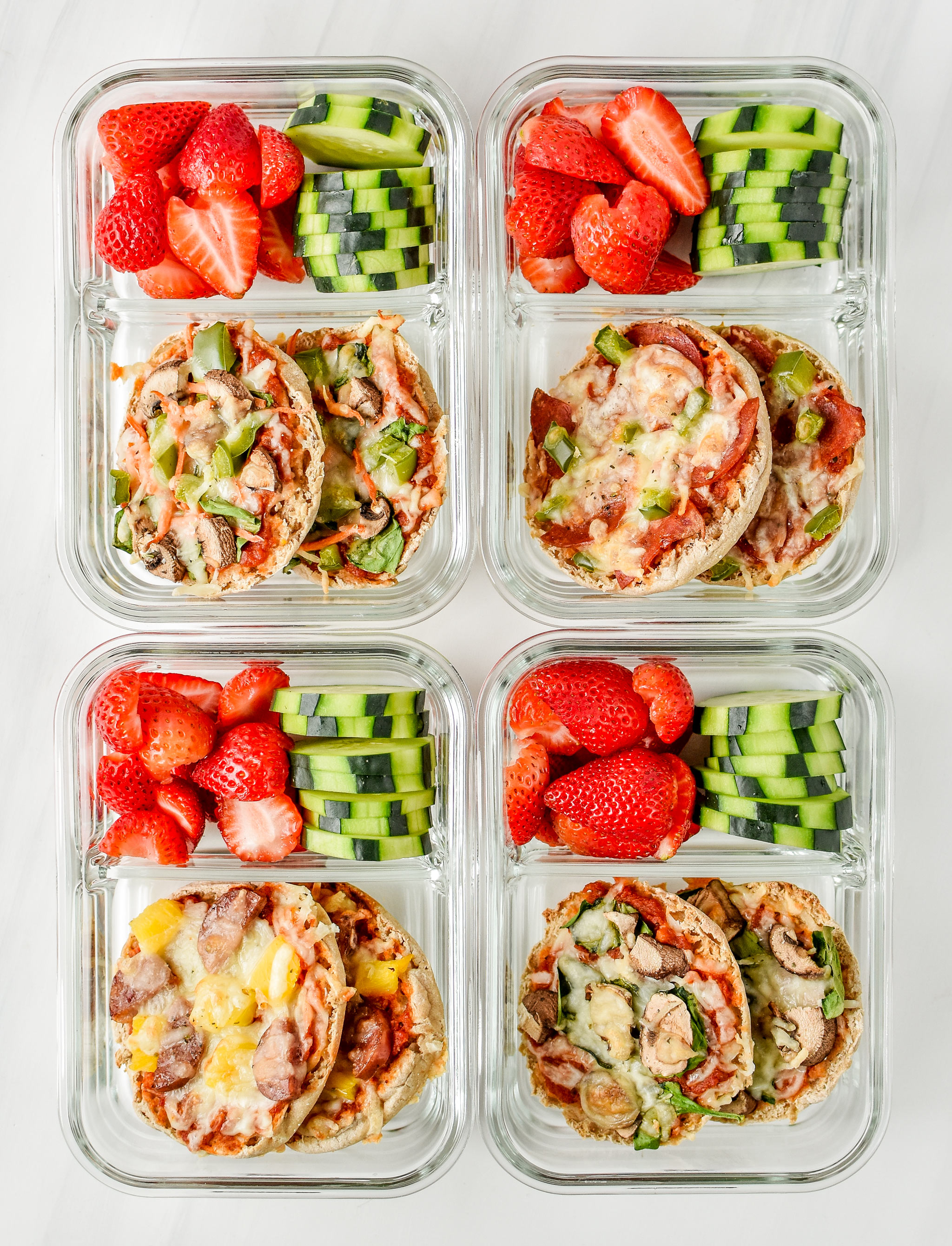 English muffin mini pizzas meal prep pictured from above in meal prep containers with fresh cut strawberries and cucumbers.