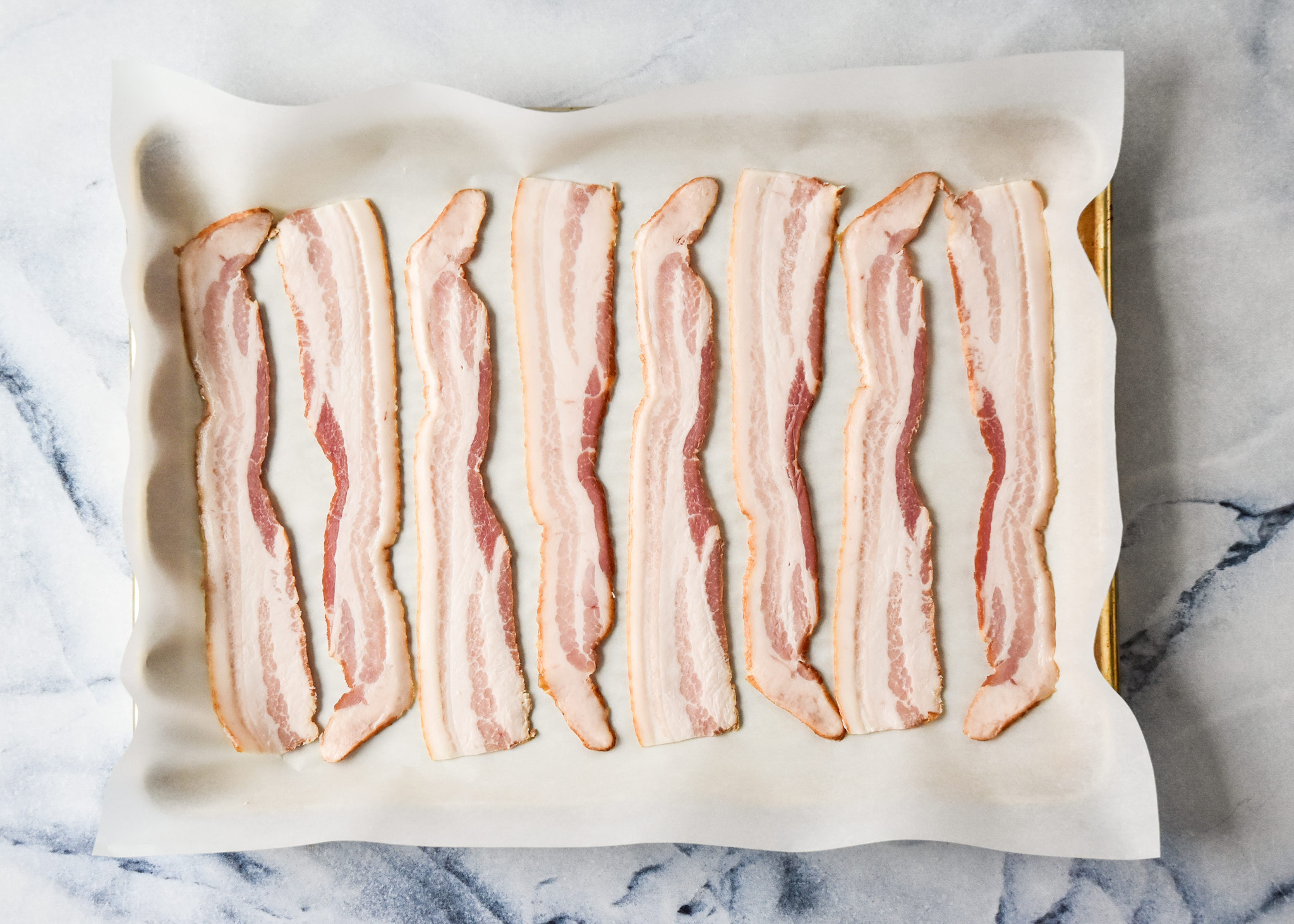 raw bacon strips on a baking sheet