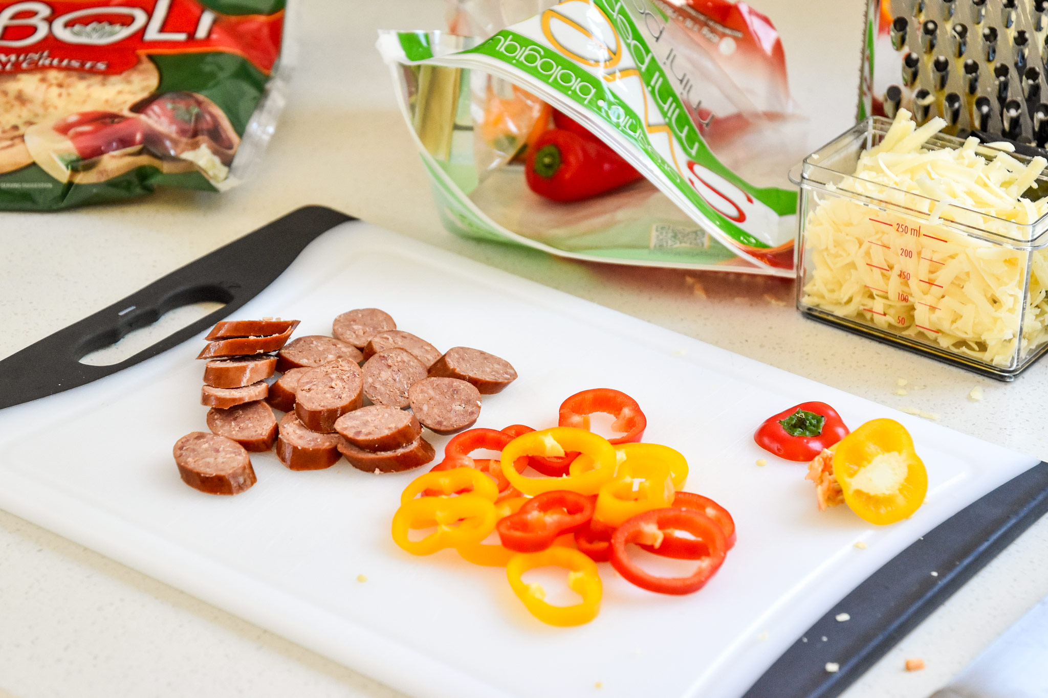 Ingredients for the sausage and pepper personal pizzas being cut on a cutting board
