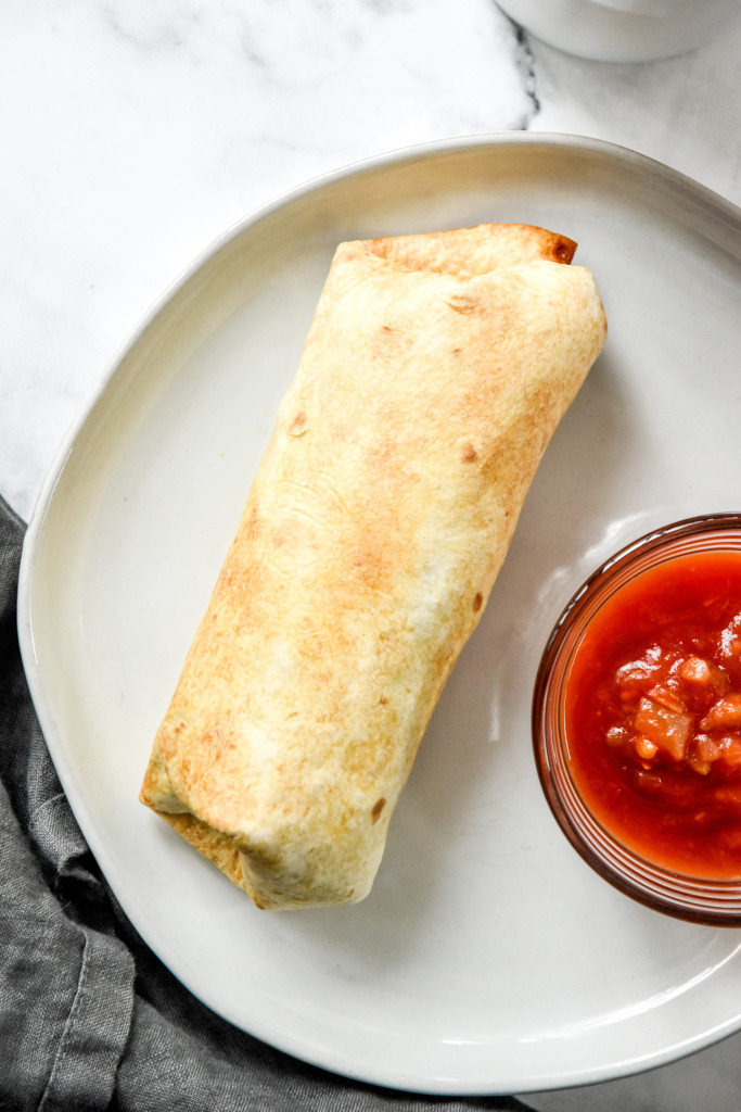 A hot browned breakfast burrito on a plate with a cup of salsa.