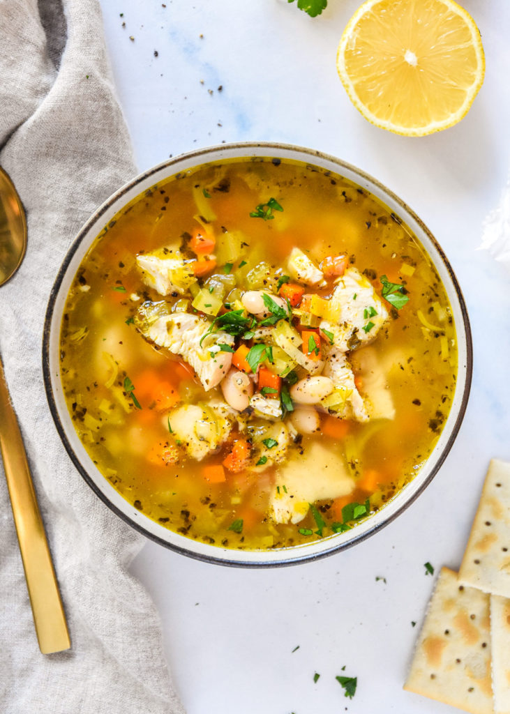 white bean lemon chicken soup in a bowl with veggies and broth, crackers and lemon on the side.