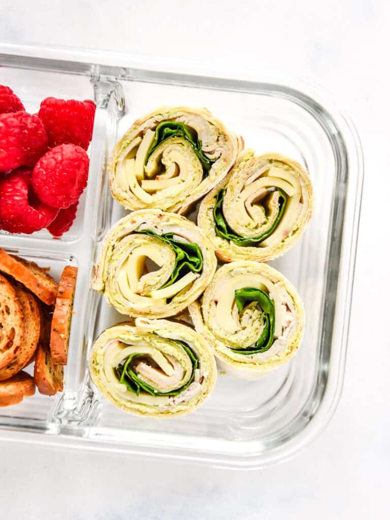 turkey pesto pinwheels in a glass meal prep container with raspberries and bagel chips.