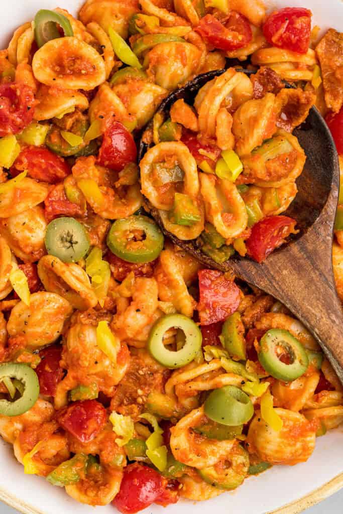 closeup of a spoonful of loaded bloody mary pasta salad in a white bowl with green olives on top.