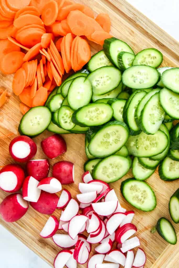 cut cucumbers, radish and carrots on a wooden cutting board.